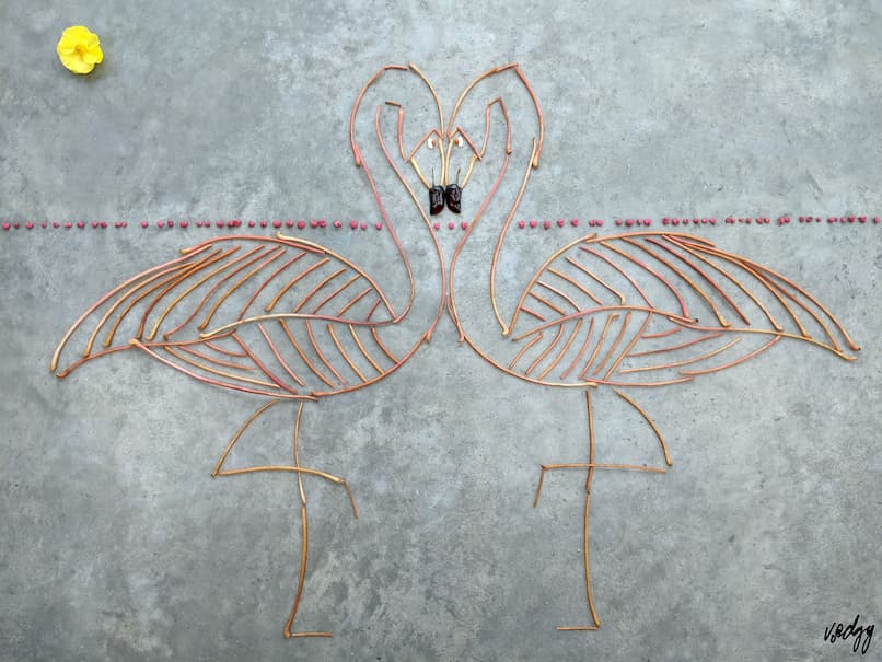 The sun (yellow ranunculus) shines over two flamingos (pink Virginia Creeper leaf stalks), each a near mirror image of the other, standing on one leg, facing each other, beaks (black chilli peppers) touching, staring into the other's eyes (white beans and orange spindleberries). Viewed from the side, their necks, heads and beaks form a heart shape. The horizon is made up of many tiny hearts (pink spindleberry pods).