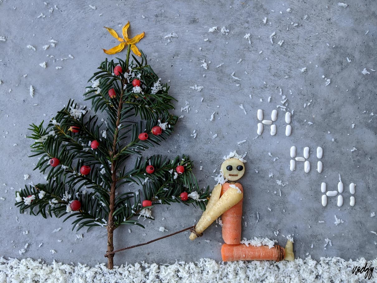 On a background of grey concrete, snow (white grated cauliflower) is falling & is on the ground. On the left a Christmas tree (sprig of yew) with baubles (red yew berries) & a star (yellow sunflower petals). To its right a letter L (orange carrot) made to look like a person with a face, arms & feet (beige carrot) is tied to the tree with a Virginia Creeper stalk. To its right are white beans spelling out HI HI HI.“
                title=